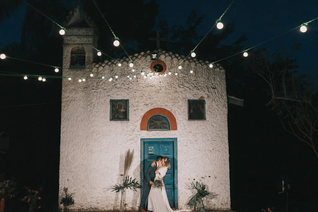 Casal de casamento em frente a uma antiga igreja branca decorada com luzes de festa à noite.