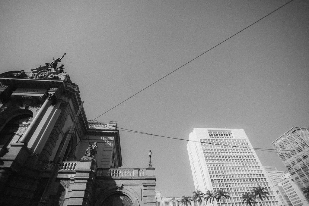 Imagem em preto e branco de prédios altos em uma cidade, com fios de energia no céu e uma arquitetura antiga e moderna