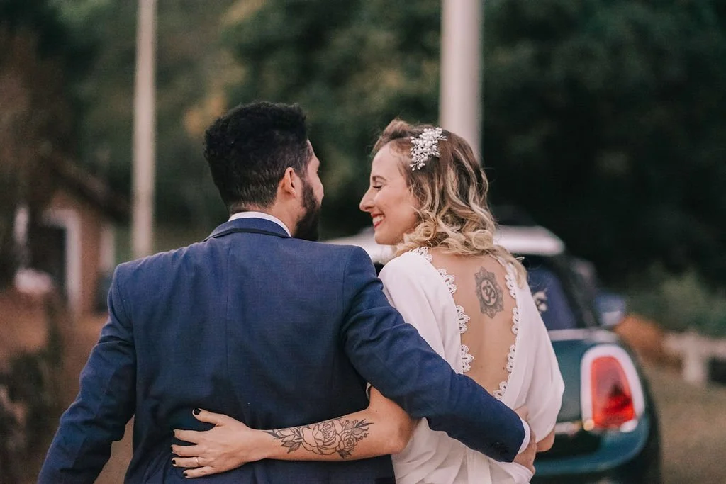Casal de noivos sorrindo e se abraçando ao ar livre com carro ao fundo, durante casamento.