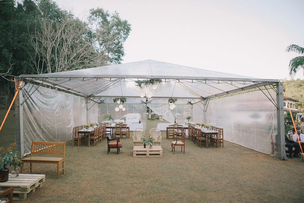 Decoração de casamento ao ar livre em uma tenda transparente com mesas e cadeiras de madeira e algumas poltronas, com árvores ao fundo.