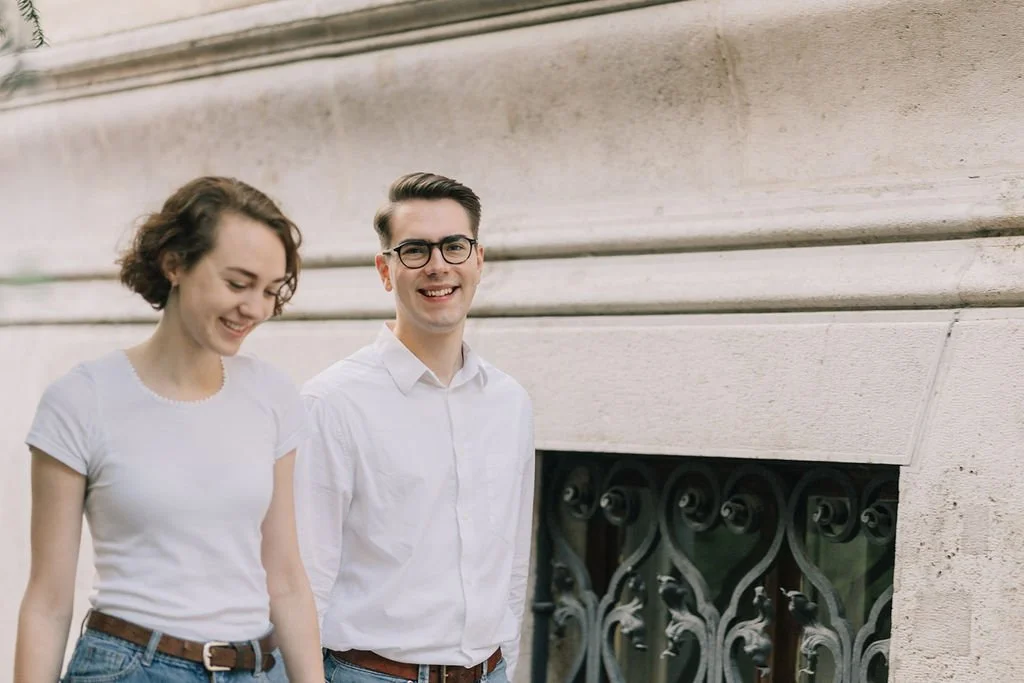 Um homem e uma mulher sorrindo, caminhando ao lado de uma parede de pedra.