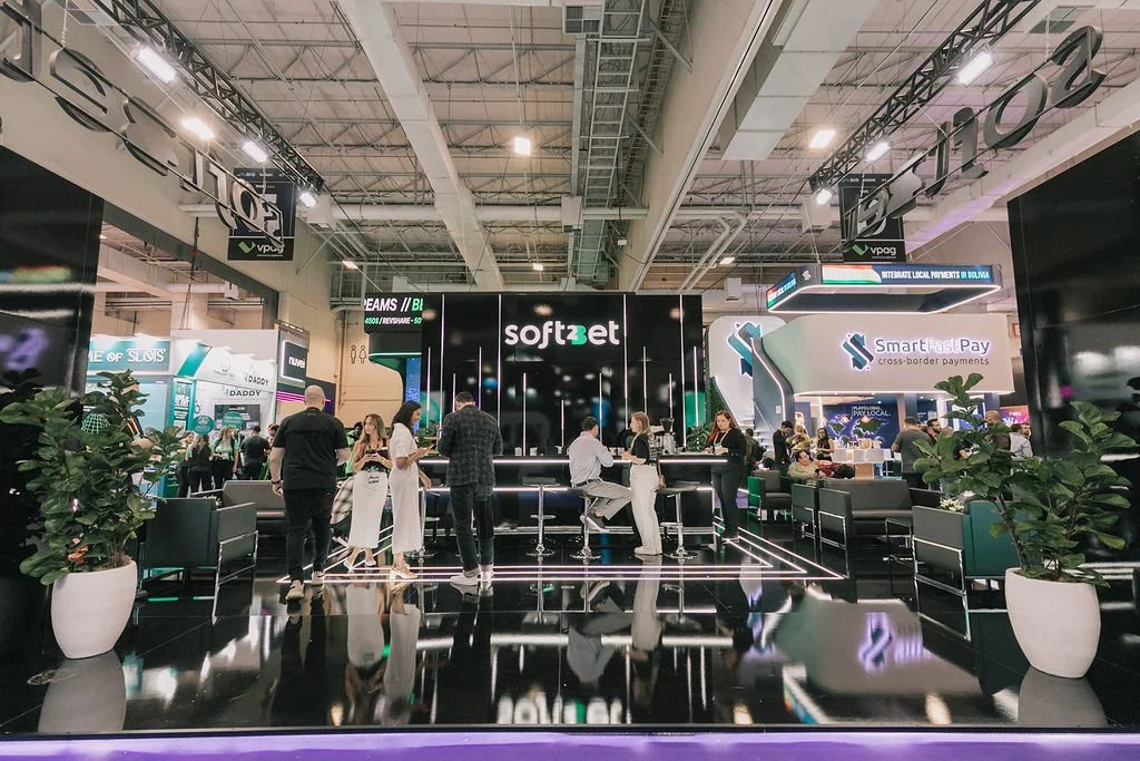 Feira de tecnologia com vários estandes, pessoas conversando e sentadas, destaque para o estande da Soft2Bet ao centro, decorado com luzes neon, e sinalização de pagamentos em várias línguas.