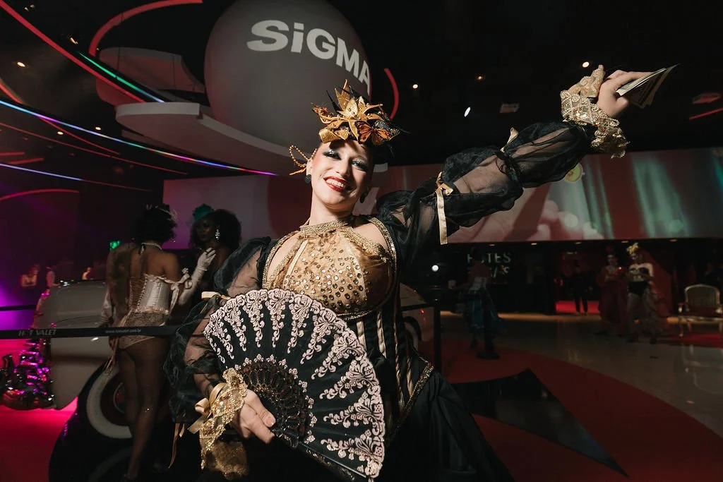 Mulher vestida com traje de época, segurando um leque decorativo, sorrindo em evento social interno com decoração moderna e pessoas ao fundo.