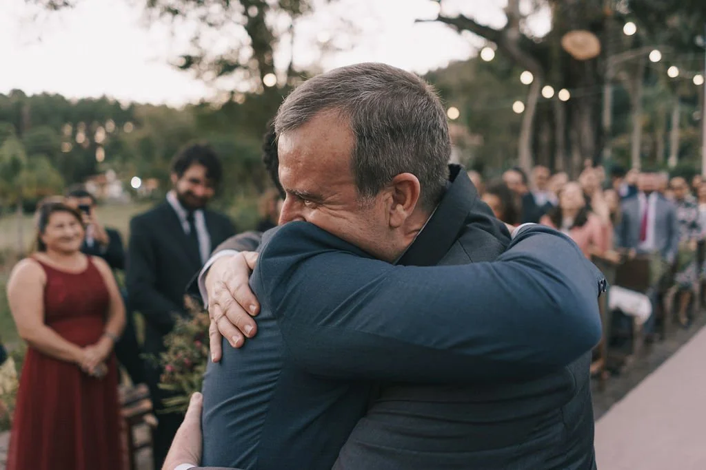 Homem com cabelo cinza e barba, vestindo terno azul, abraçando alguém em evento ao ar livre, com várias pessoas assistindo ao fundo.