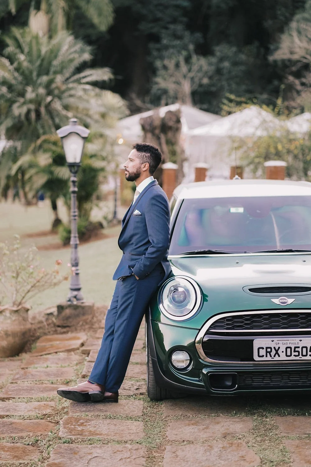 Homem de terno azul escuro apoiado no carro Mini Cooper verde escuro, em um ambiente ao ar livre com árvores e luminária antiga ao fundo.
