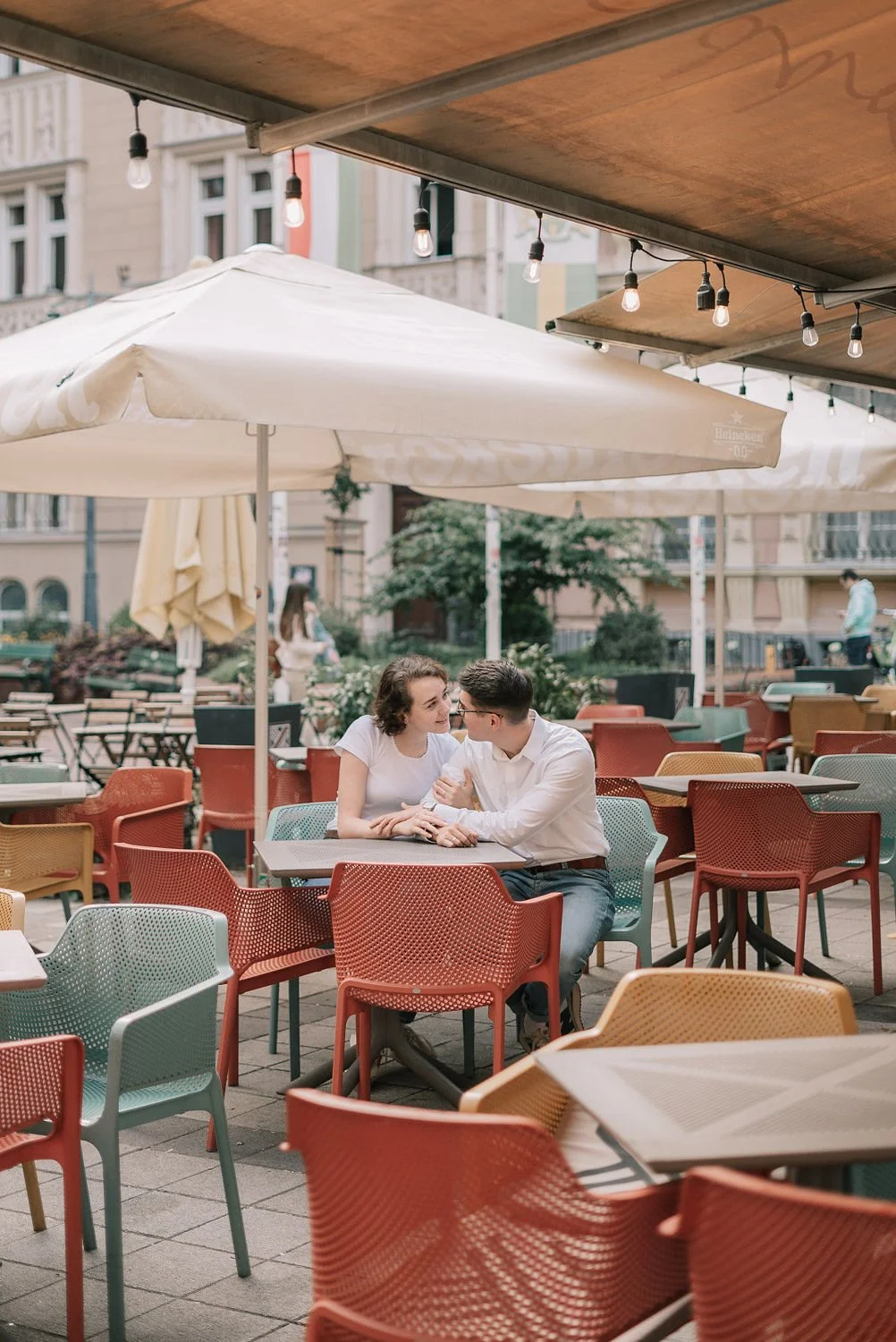 Casal sentado em mesa ao ar livre em um café, conversando sob guarda-sóis, com iluminação de luzes penduradas e edifícios ao fundo.