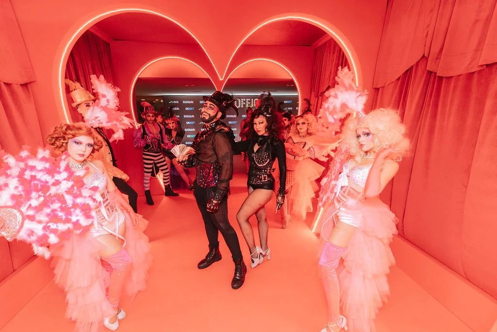 Grupo de pessoas vestidas com fantasias de Carnaval em um cenário iluminado com luzes rosa e fundo com coração neon.