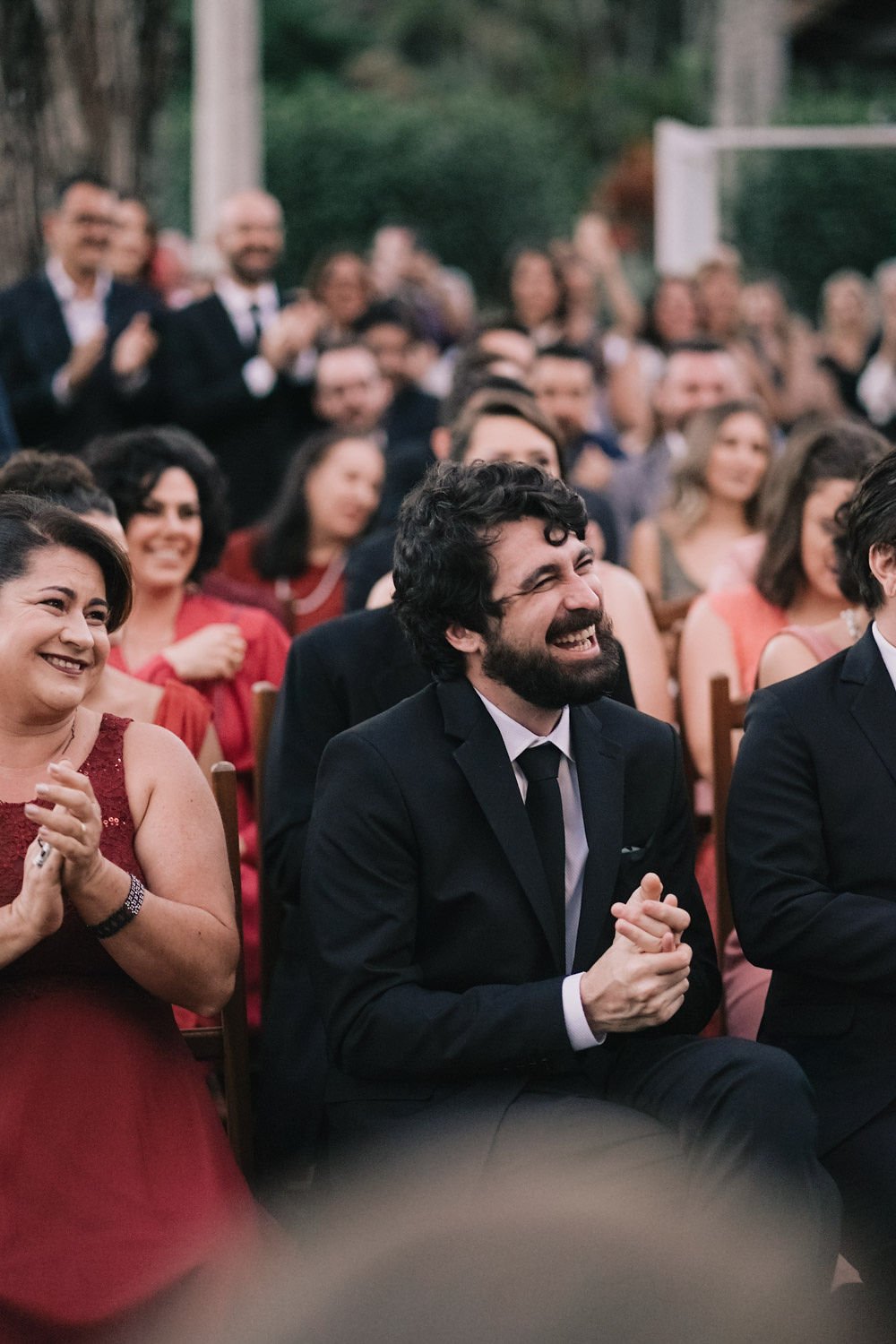 Grupo de pessoas rindo e aplaudindo em um evento ao ar livre na natureza, vestidas de formal, com destaque para um homem de barba e cabelo cacheado ao centro, usando terno preto, sorrindo.