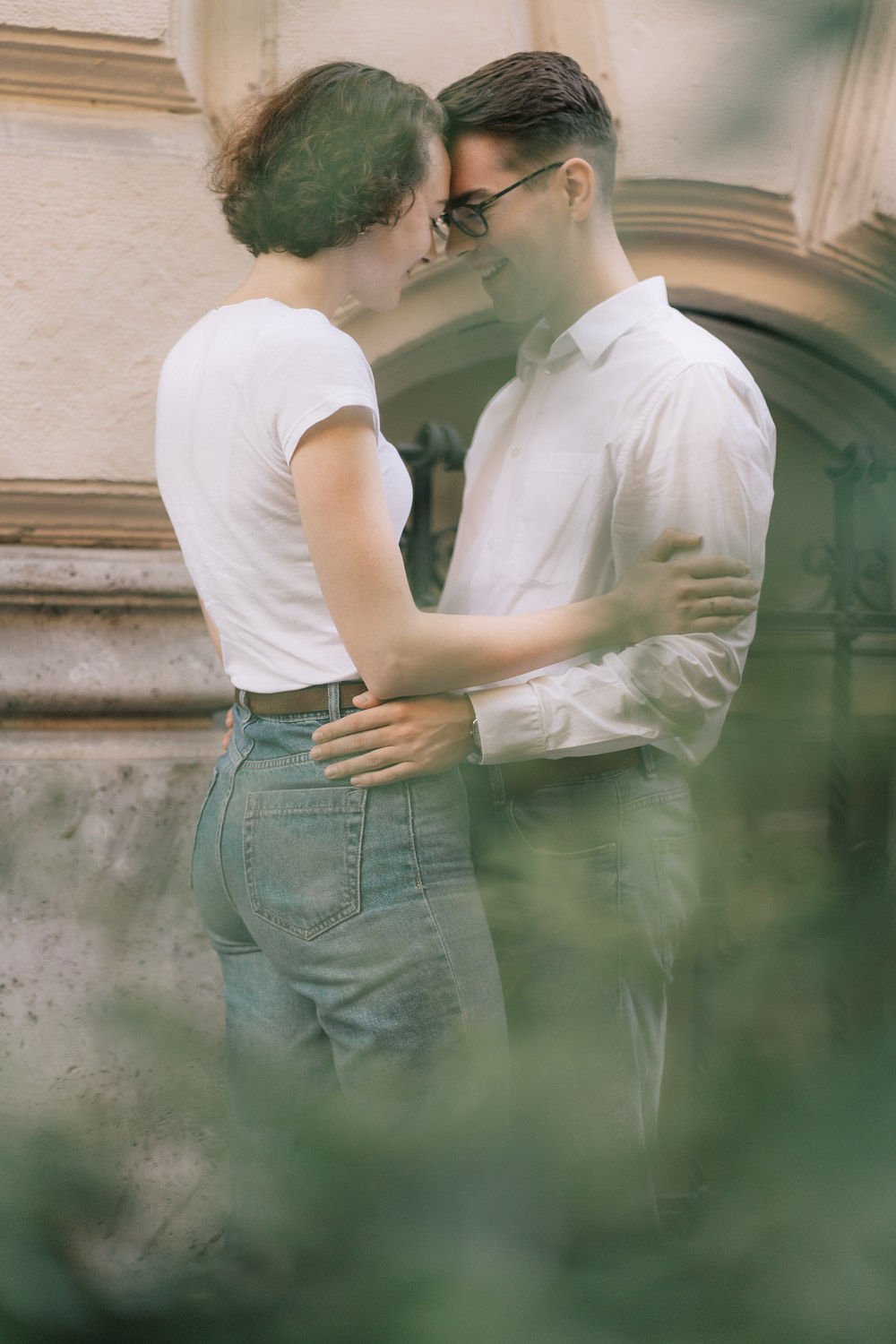Um casal jovem, sorrindo, se abraçando de cabeça inclinada, em um ambiente externo com parede de pedra ao fundo e plantas borradas ao redor.