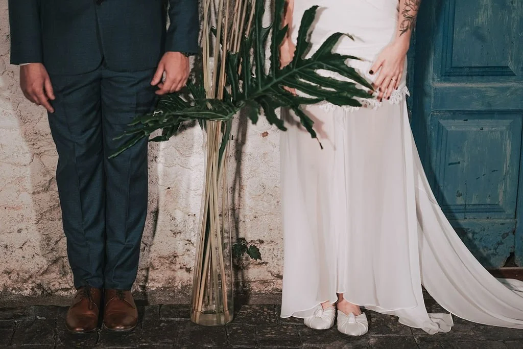 Parte inferior de um homem vestido com terno azul e sapatos marrons, ao lado de uma mulher com vestido branco de casamento e sapatos brancos, segurando um grande arranjo de plantas verdes na frente de uma parede de pedra e uma porta azul envelhecida.