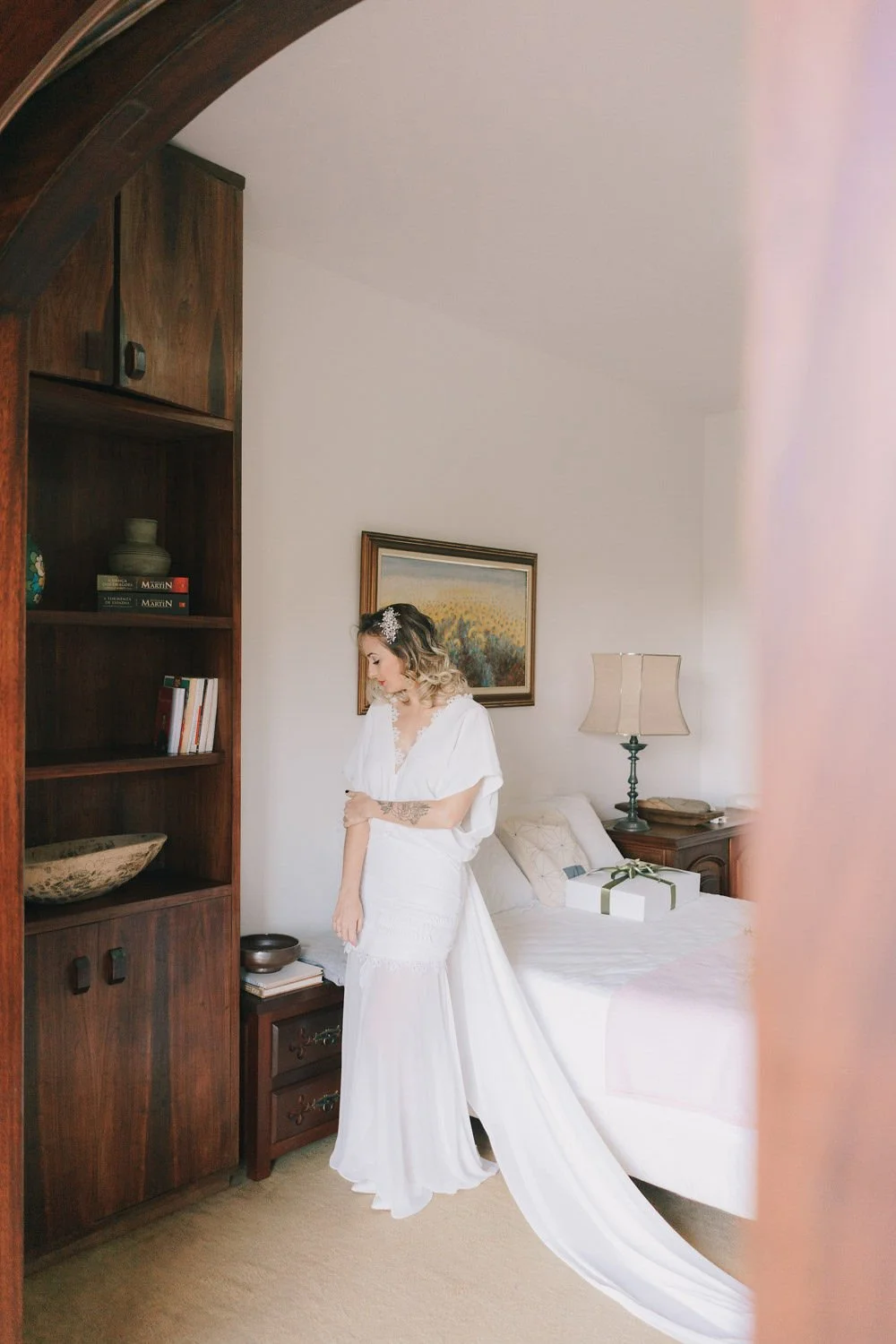 Mulher vestida com vestido branco de noiva em um quarto, ao lado da cama com uma caixa de presente.