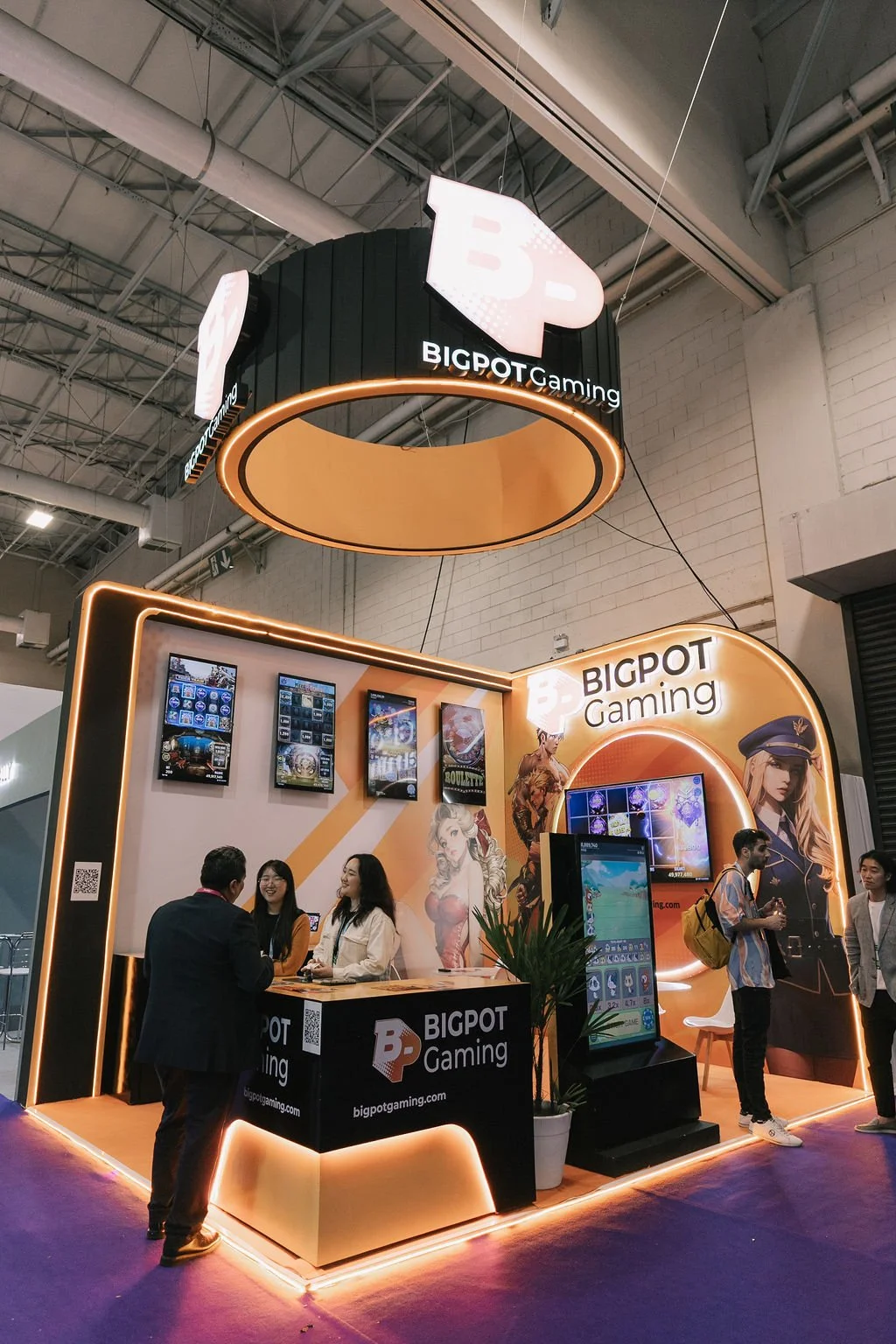 Stande de uma feira de jogos da BIGPOT Gaming com várias telas de jogos digitais e pessoas interagindo, decorado com iluminação de led e arte de personagens de jogos.