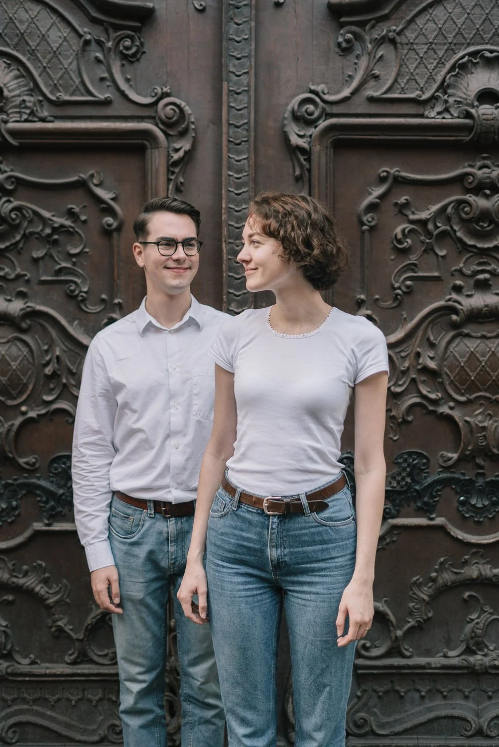 Um homem de óculos e uma mulher com cabelo cacheado diante de uma porta de madeira ornamental, ambos usando roupas casuais brancas e jeans.