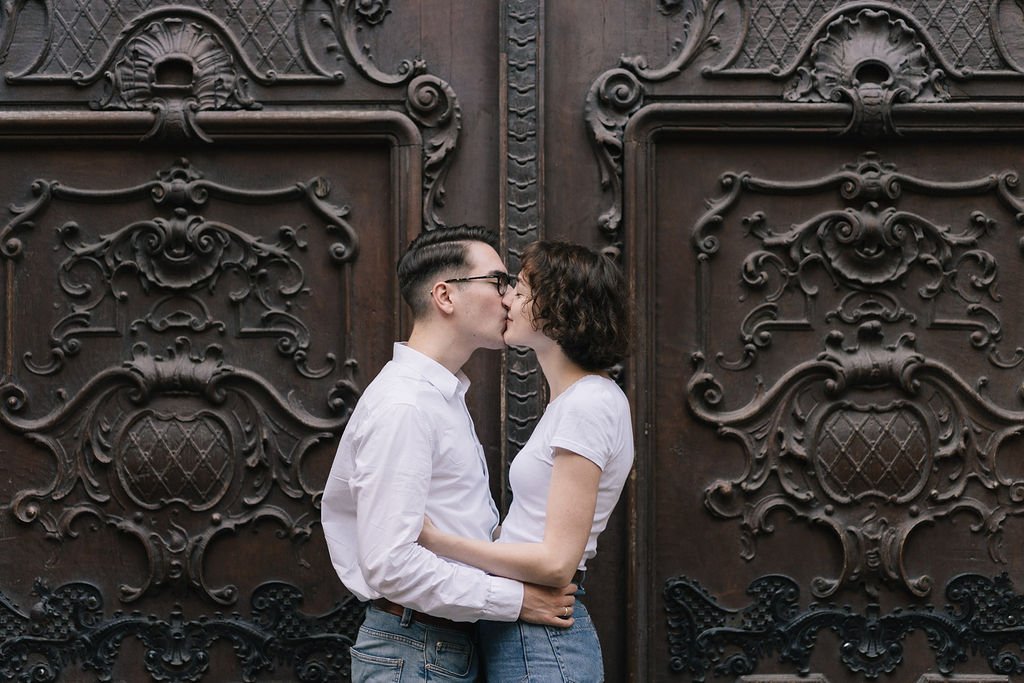 Um casal se beijando na rua, diante de uma porta de ferro decorada com detalhes elaborados.