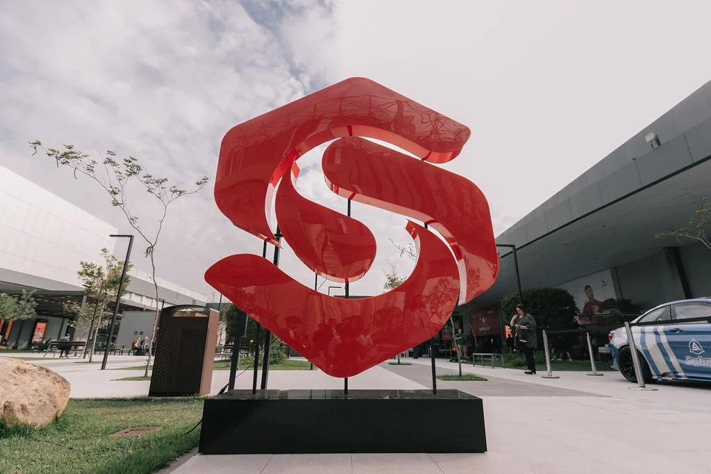 Escultura grande de um símbolo estilizado em cor vermelha, em um espaço ao ar livre em frente a edifícios comerciais, com algumas pessoas ao redor.