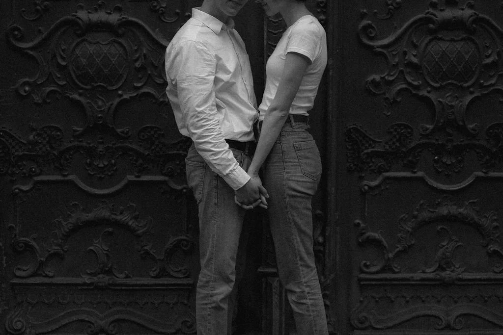 Homem e mulher de mãos dadas, de pé perto de uma porta de ferro decorada, em preto e branco.