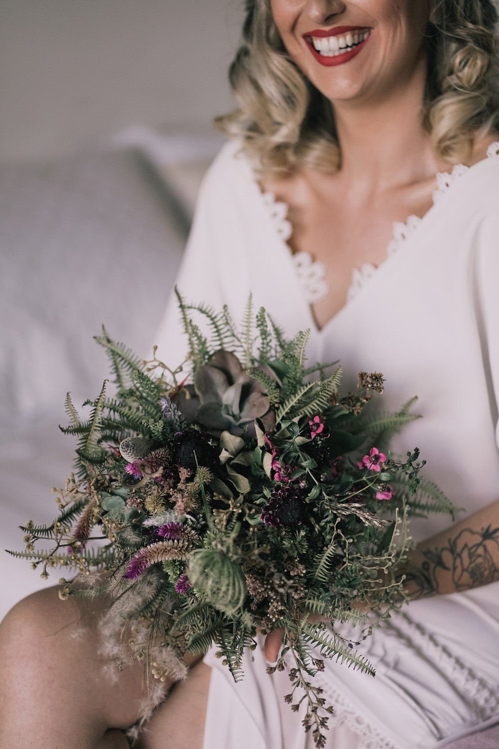 Mulher sorridente com cabelo cacheado, vestida de branco, segurando um buquê de flores variadas com folhagens verdes e detalhes em rosa e roxo.
