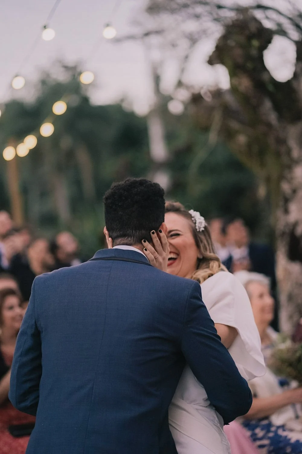 Casal se beijando em festa ao ar livre, com pessoas ao fundo e luzes decorativas.