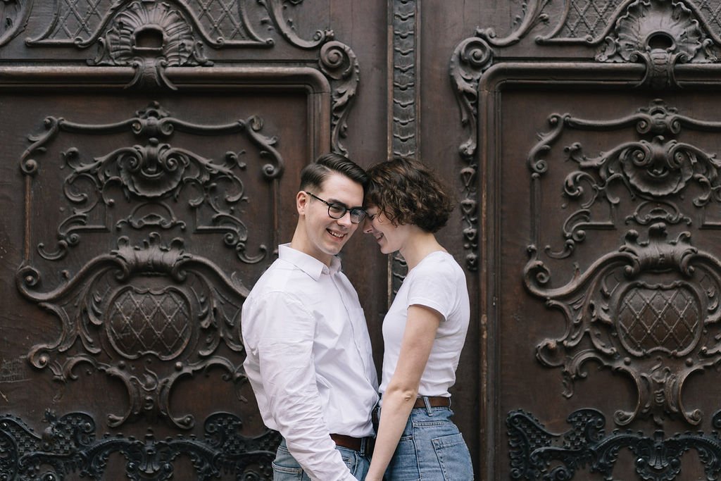 Casal jovem rindo e sorrindo juntos em frente a uma porta de madeira ornamentada.