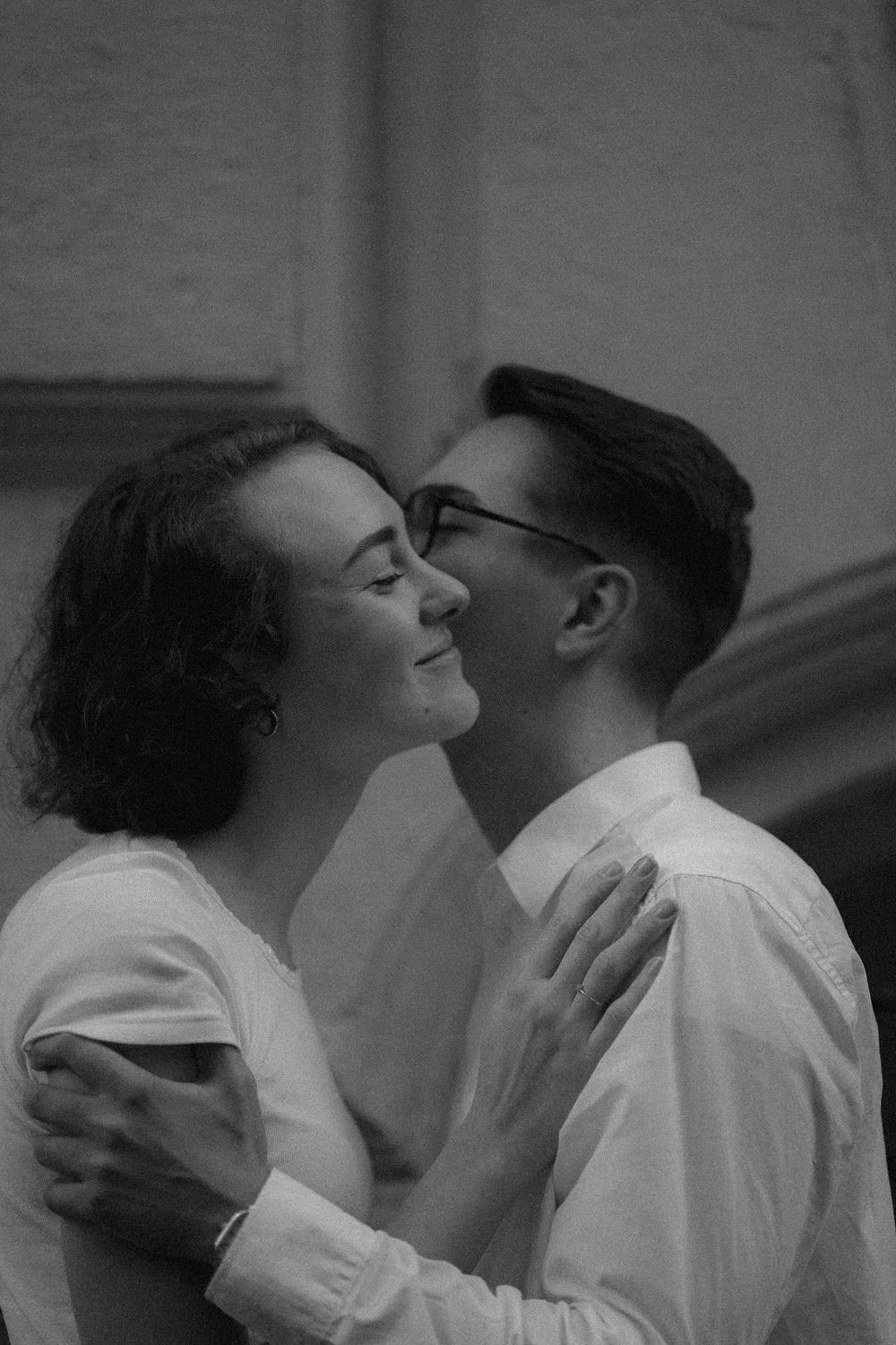 Um casal sorridente, de pele clara, se beija carinhosamente, com ela segurando o ombro do rapaz, em um ambiente interno, em preto e branco.