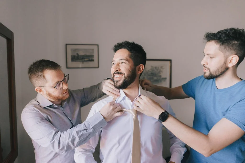 Três homens ajudando um homem a colocar uma gravata em um ambiente interno.