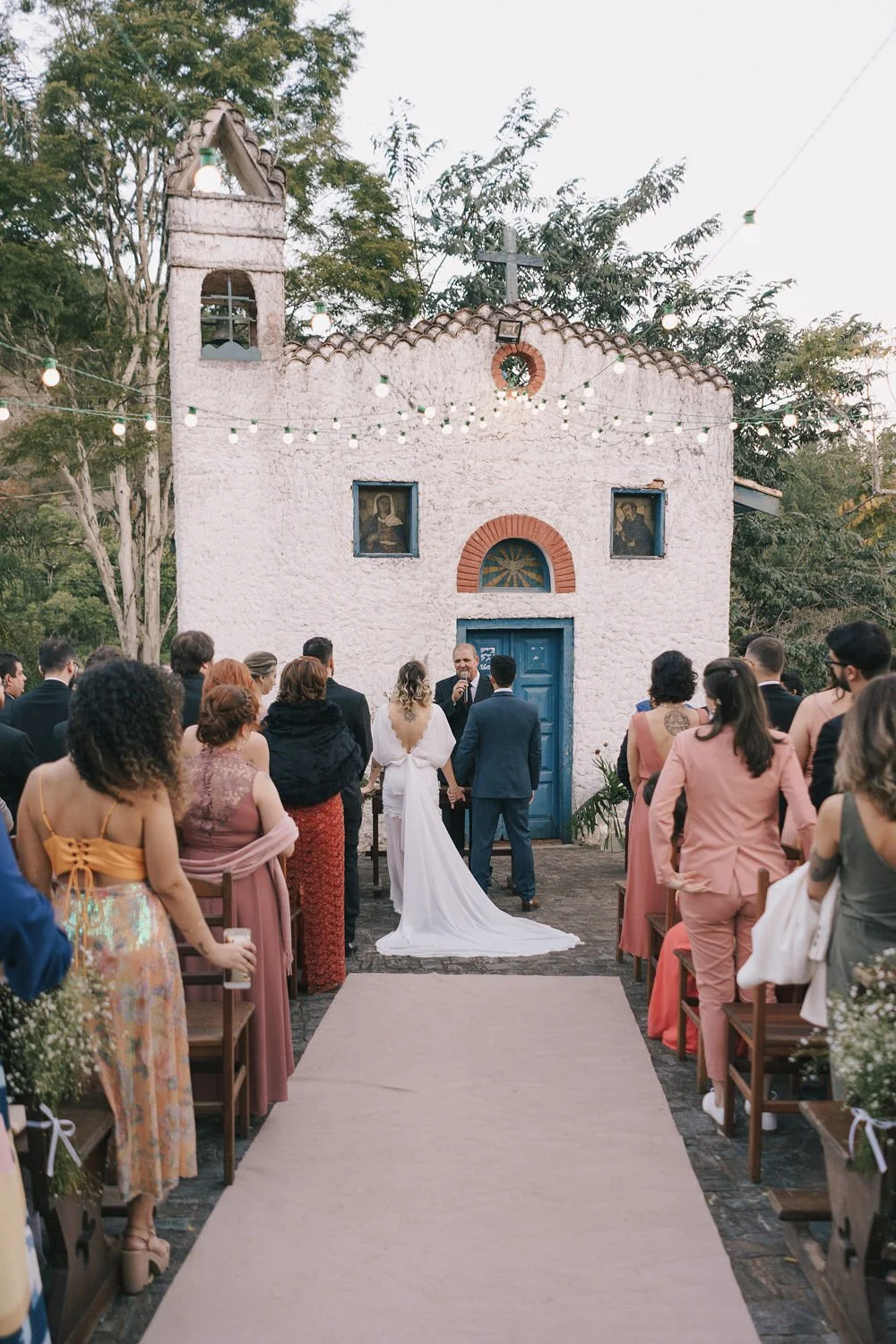 Casamento ao ar livre com noivas e convidados diante de uma pequena igreja branca com porta azul, árvores ao redor e iluminação de luzes penduradas.