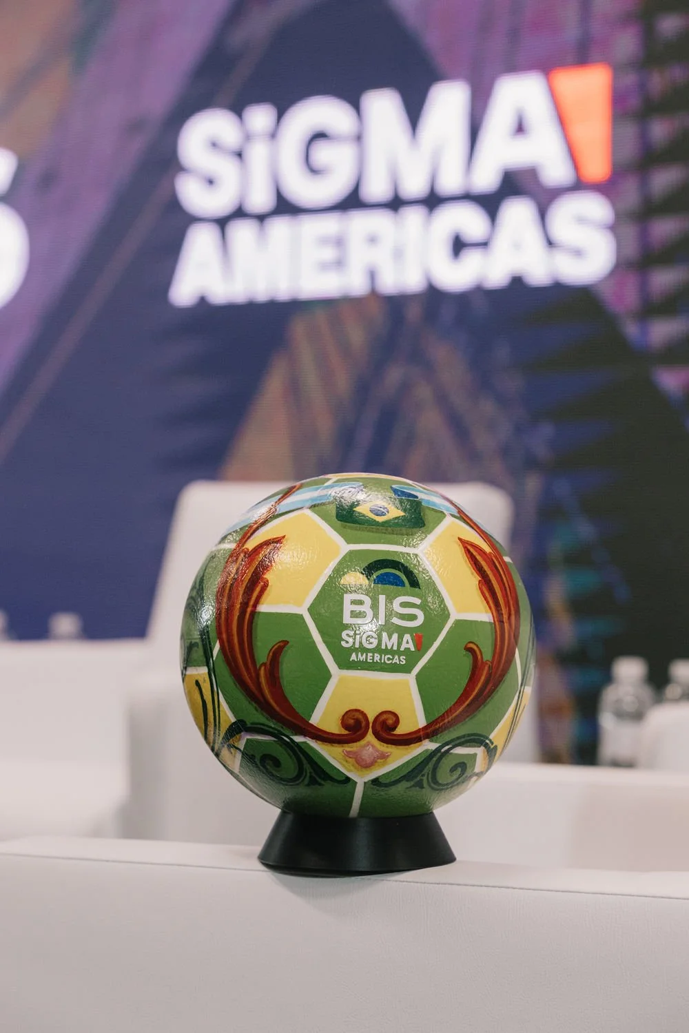 Sala de conferências com uma bola de futebol decorativa na frente, com o logotipo de Bisigma Américas. Fundo com uma faixa de banner azul com o texto parcialmente visível 'SIGMA AMERICAS'.