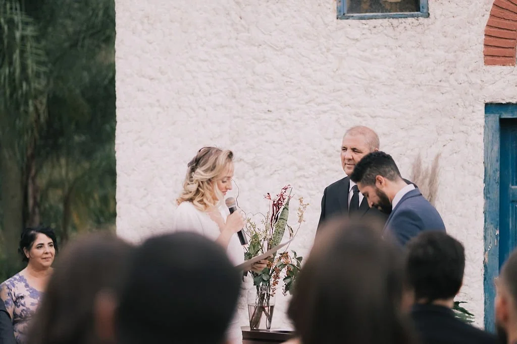 Casamento ao ar livre com uma mulher lendo votos enquanto outro homem ouve, com convidados ao redor