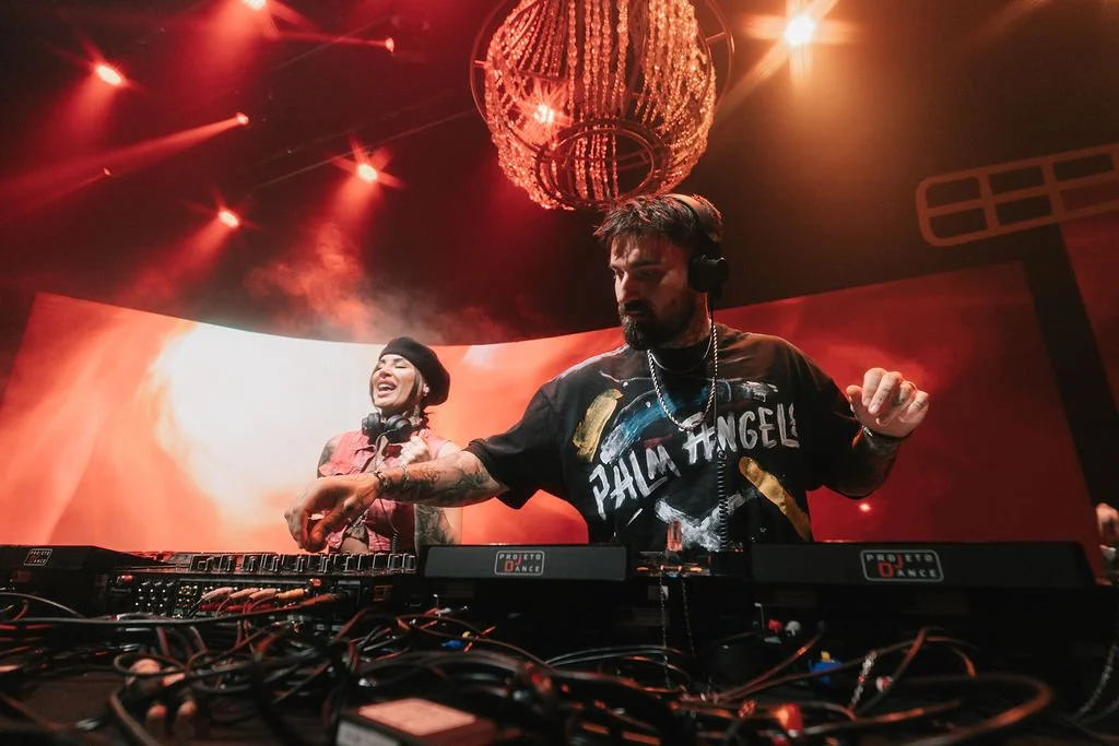 Dois DJs se apresentam em uma pista de dança, com iluminação vermelha e um cristal de lustre acima, um deles tem barba, camiseta preta com o nome 'Palm Angels' e usa fone de ouvido, enquanto a outra mulher sorri ao fundo, também usando fone de ouvido
