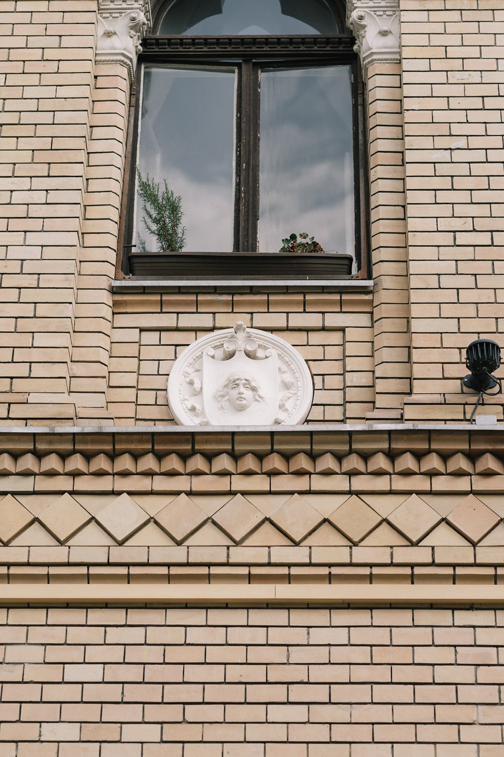 Detalhe de uma fachada de edifício de alvenaria com uma janela de vidro e uma decoração acima dela com uma escultura de uma cabeça de criança com cabelo desgrenhado, em relevo, em moldura oval branca.