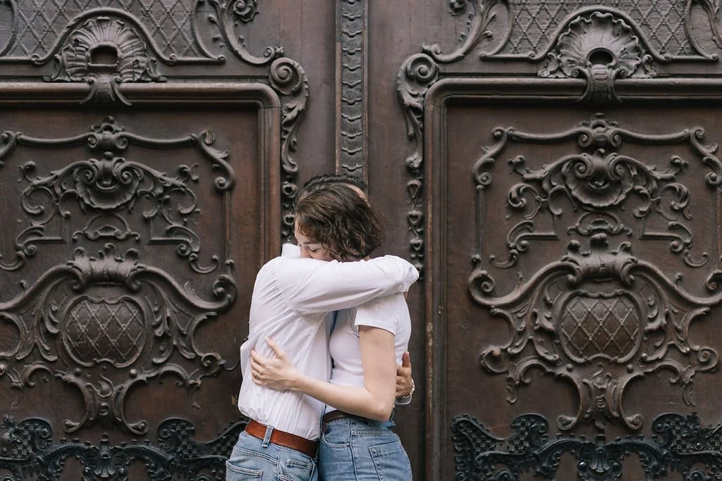 Duas pessoas abraçadas perto de uma porta de madeira decorada com detalhes elaborados.