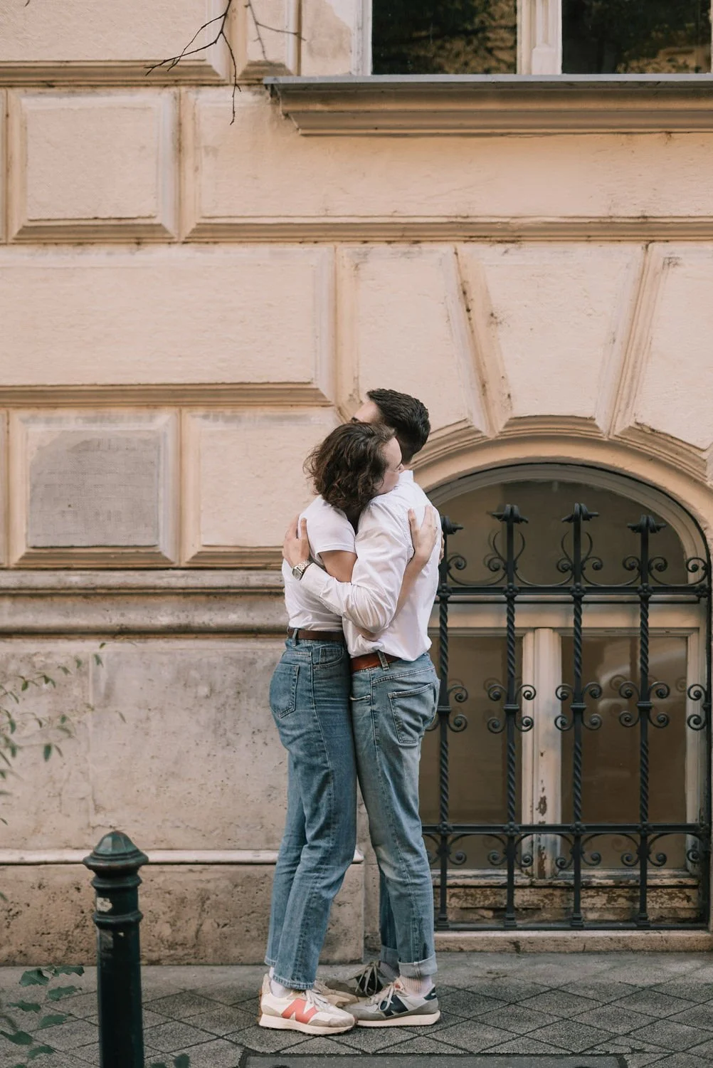 Casal se abraçando na rua, com fundo de parede de prédio antigo com janela e grade de ferro.