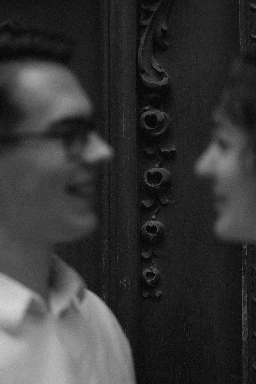 Imagem em preto e branco de duas pessoas com sotaque brasileiro, uma de perfil e outra de frente, olhando uma para a outra, com uma parede ao fundo que possui uma decoração de objetos circulares de madeira empilhados verticalmente.