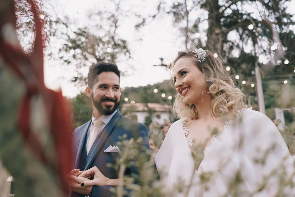Noivos em casamento ao ar livre, sorrindo um para o outro, com luzes decorativas e árvores ao fundo.
