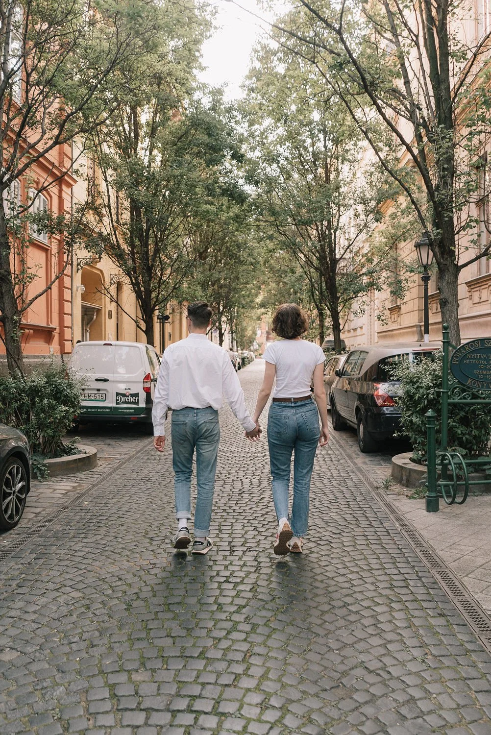 Casal caminhando de mãos dadas pela rua de paralelepípedos em uma área residencial, cercada por árvores e carros estacionados.