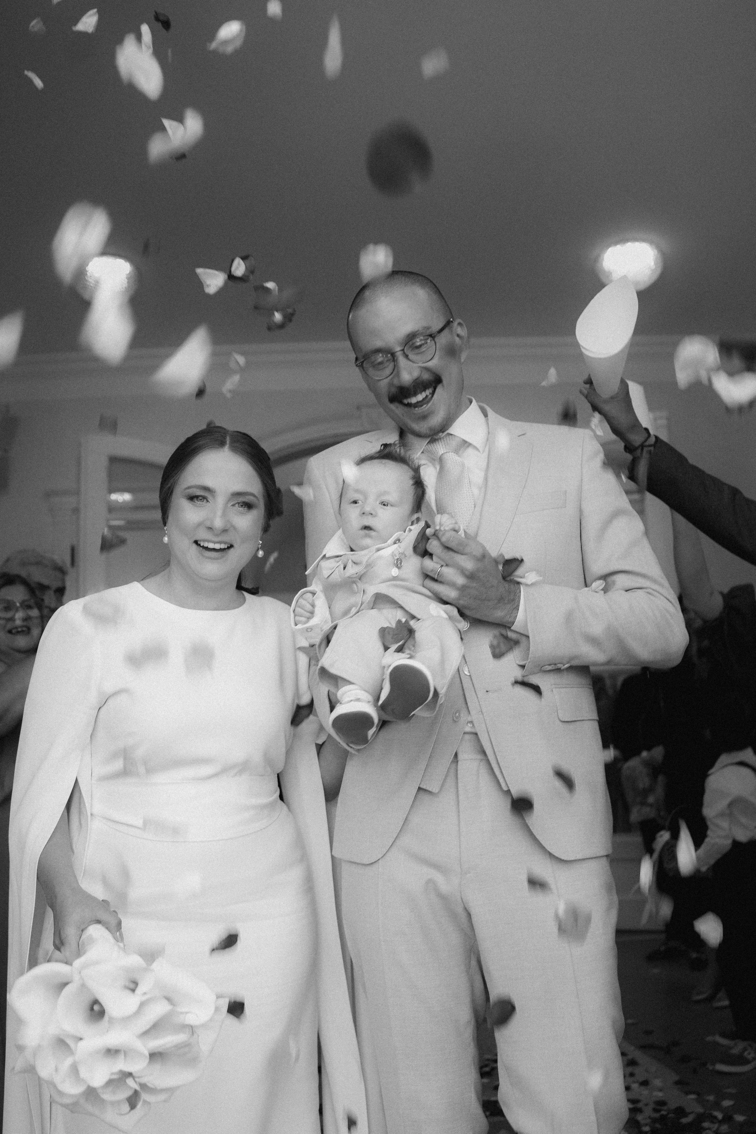 Casal recém-casado comemorando com um bebê, enquanto pétalas de flores caem, em uma celebração de casamento.