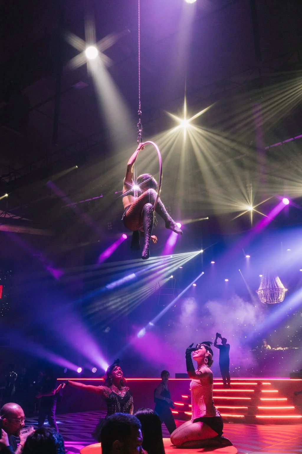 Apresentação de circus com uma pessoa suspensa em uma argola no teto, iluminada por luzes coloridas, enquanto outras pessoas estão no palco, algumas de joelhos, em ambiente de show com iluminação vibrante e elementos de palco.
