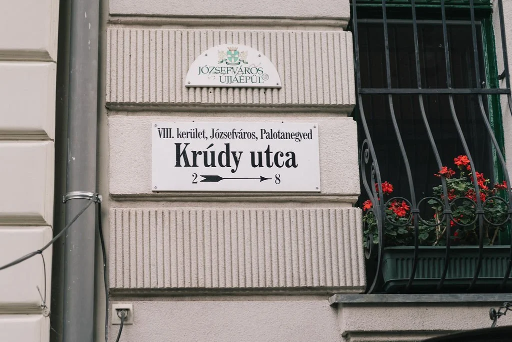 Placa de rua com o nome "Krudy utca" e referências a Józsefváros, Budapest, com ornamentos e flores na janela ao lado.