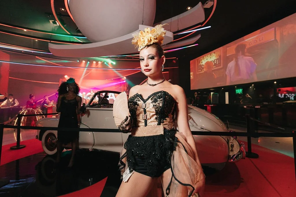 Mulher vestida com traje extravagante de festa, com cabelo preso e acessório de cabeça decorativo, posando na frente de um carro clássico branco em uma exposição de vidro, com um ambiente de luzes coloridas e pessoas ao fundo.
