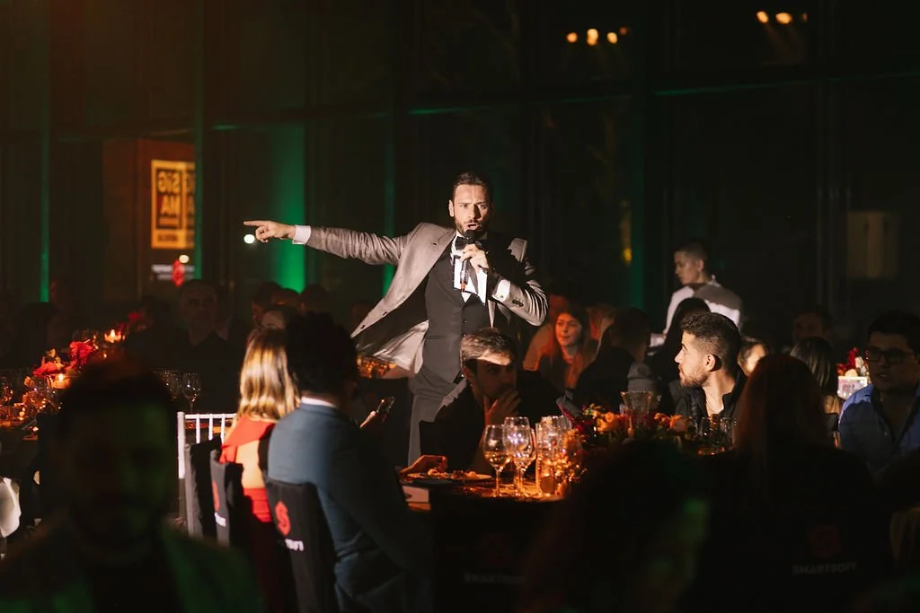 Homem em terno escuro faz discurso ou apresentação com microfone em evento formal, enquanto público assiste sentado a mesas decoradas com velas e copos de vidro.