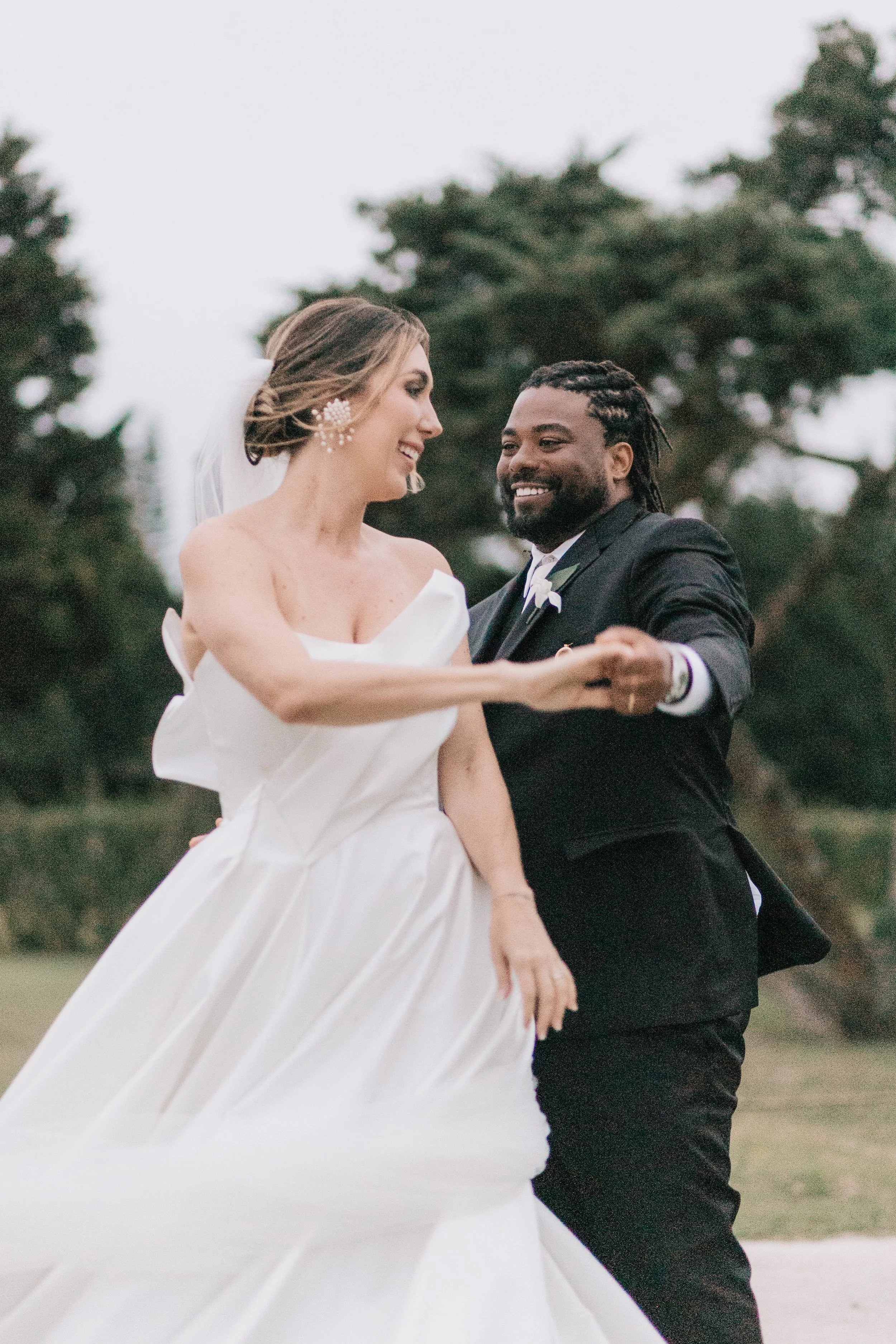Casal de noivos dançando ao ar livre, a noiva usa vestido branco e o noivo terno preto, em um cenário natural com árvores ao fundo.