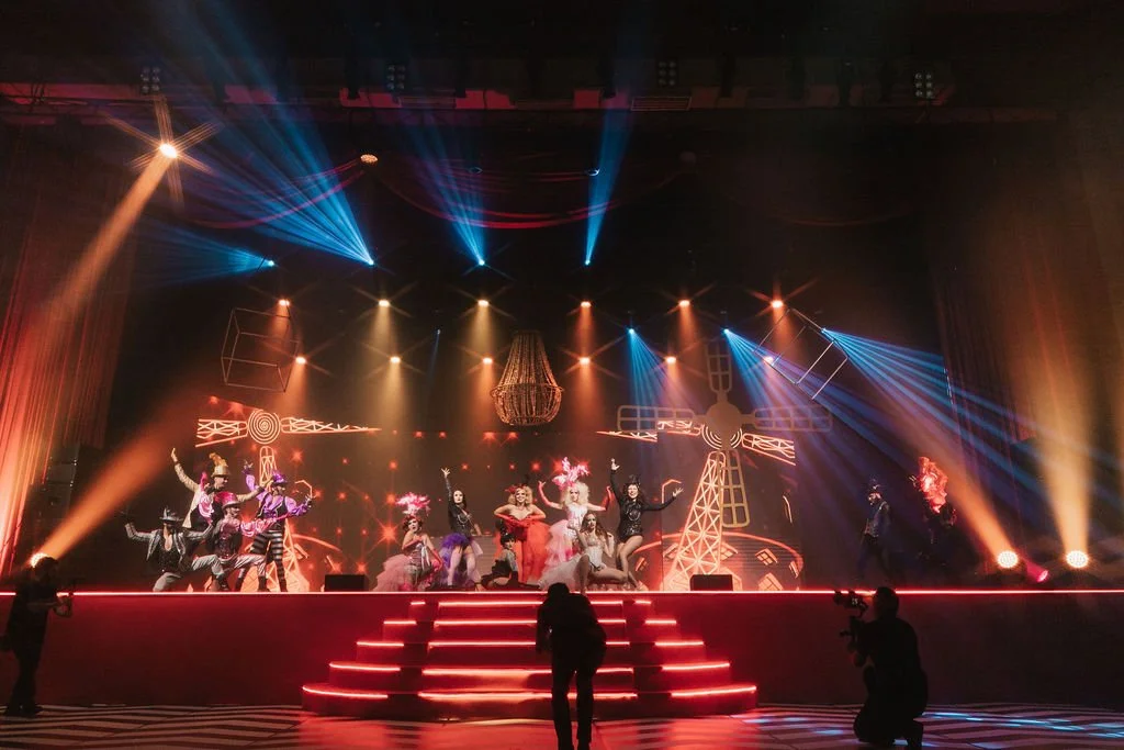 Show de dança com performers em fantasias coloridas, iluminação de palco vibrante e efeito de luzes no cenário de teatro.