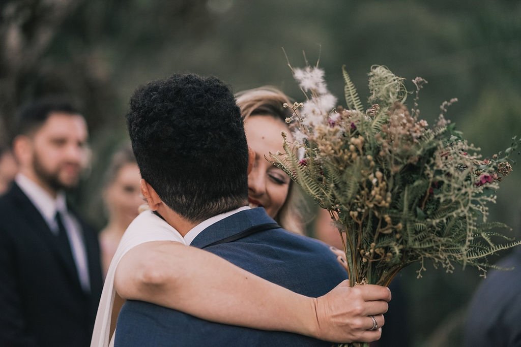 Casal de casamento se abraçando, mulher chorando de felicidade, segurando um buquê de flores. Pessoas ao fundo observando.