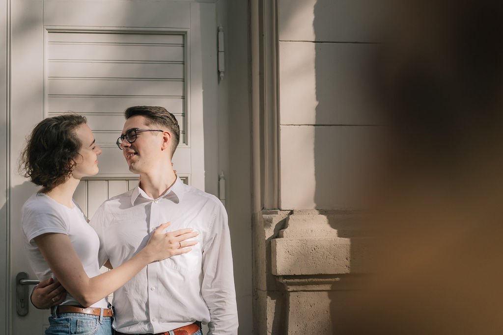 Um casal sorridente formando um casal na rua, com um fundo de parede de cimento e uma porta branca, ambos olhando um no outro e em um momento de carinho.