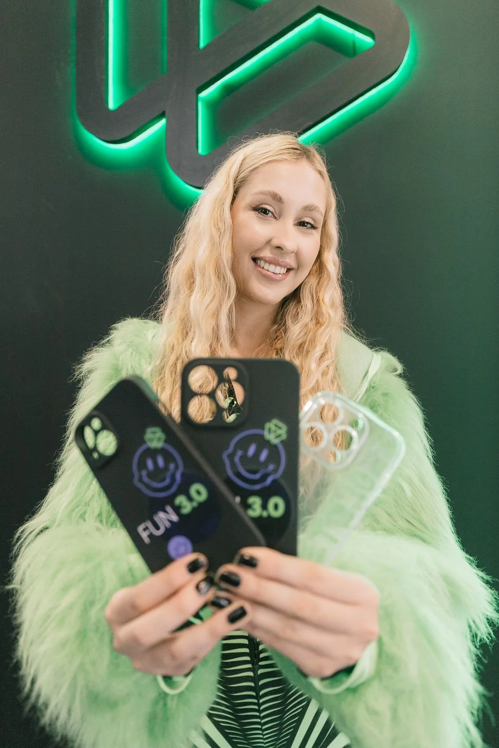 Mulher sorridente com cabelo loiro cacheado segurando três capas de celular coloridas com desenhos de smiley e a palavra 'FUN 3.0', em um fundo escuro com iluminação verde