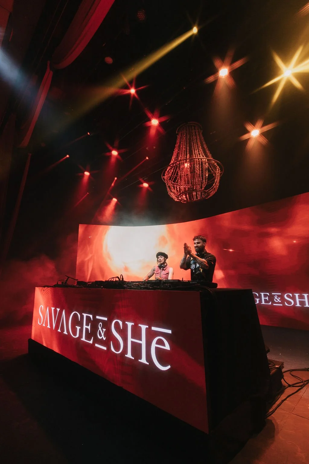 DJs performing on stage with a large LED screen displaying an orange background, red and yellow stage lights, and an elegant chandelier hanging from the ceiling. The DJ booth has a sign with the words "SAVAGE & SHE".