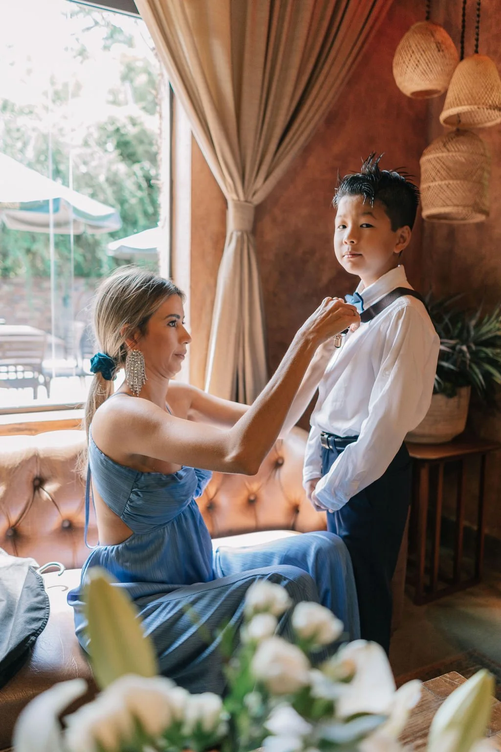 Um mulher ajudando um menino a ajustar a gravata, dentro de um ambiente bem decorado com cortinas e luminárias de macramê.