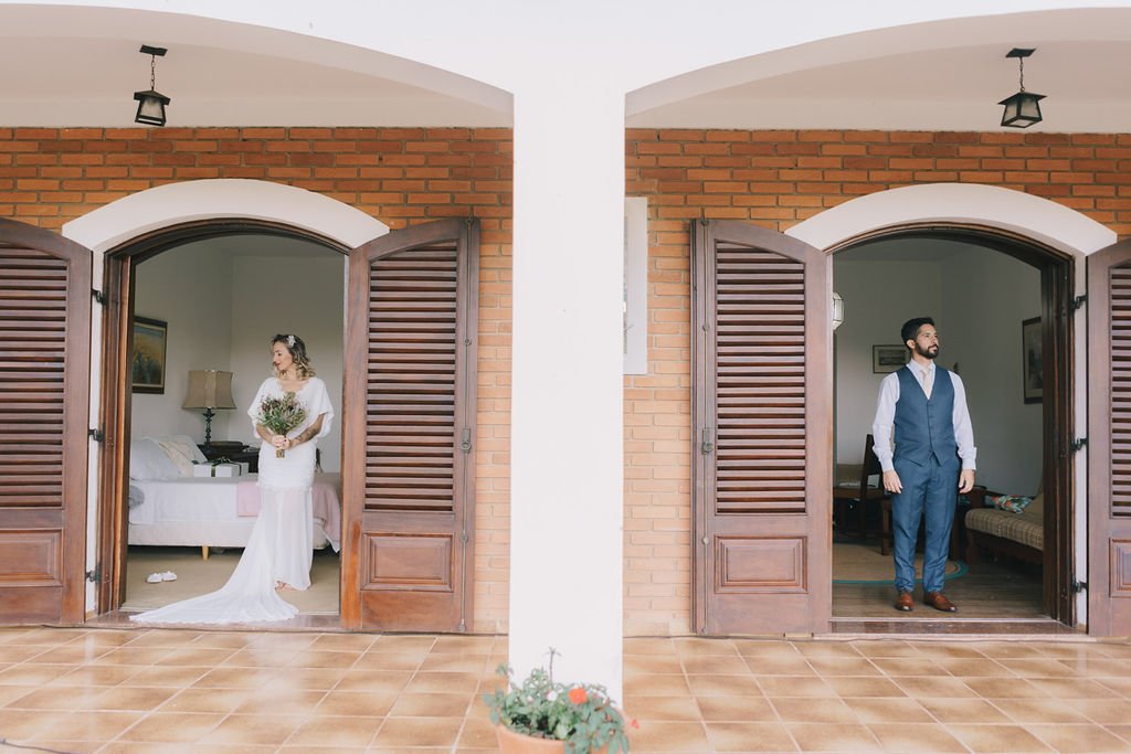 Casal de noivos separado por uma parede, cada um em uma porta de uma casa com portas de madeira. A noiva está vestida de branco, segurando um buquê de flores, dentro da casa à esquerda. O noivo está de terno azul escuro, com camisas branca, dentro da