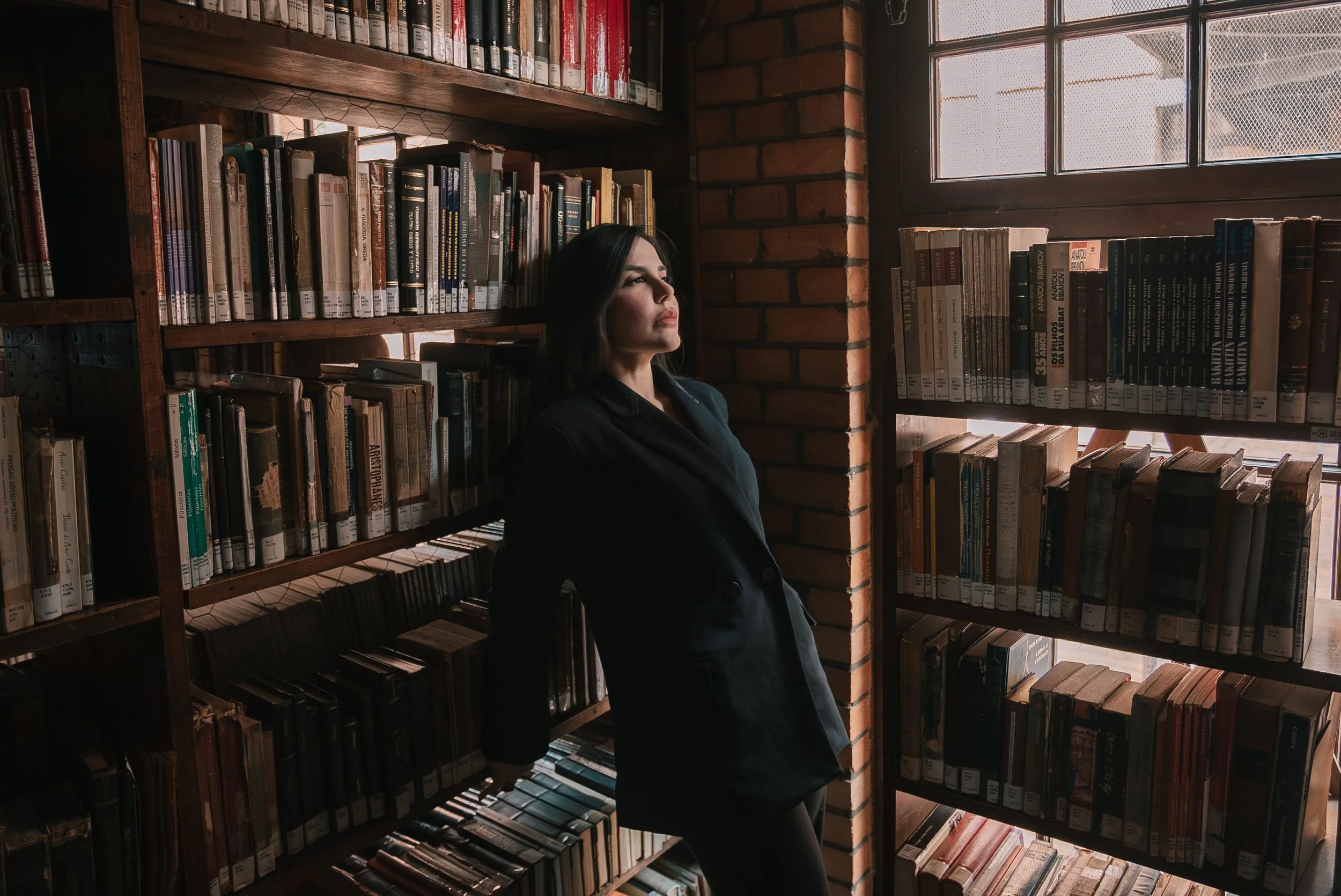 Mulher de cabelo escuro e de blazer preto apoiada em uma estante de livros na biblioteca, com luz natural entrando por uma janela ao lado.