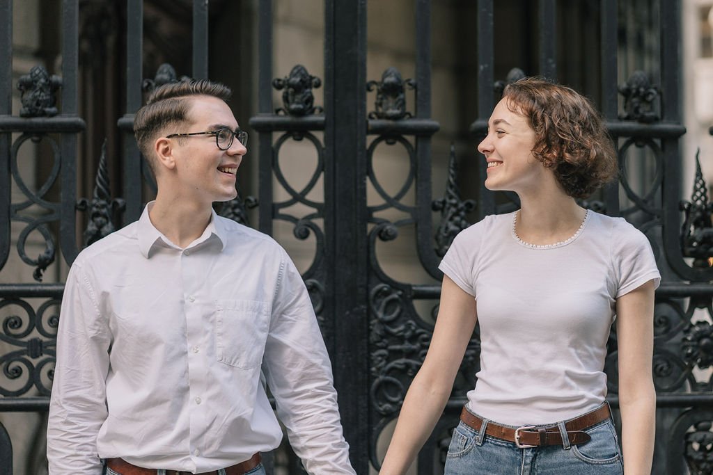 Casal jovem sorrindo e segurando as mãos na frente de uma grade de ferro preta.